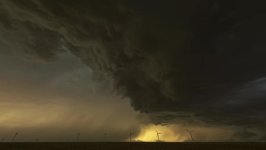 A huge storm cloud with lightning falling passing over a field with windmills