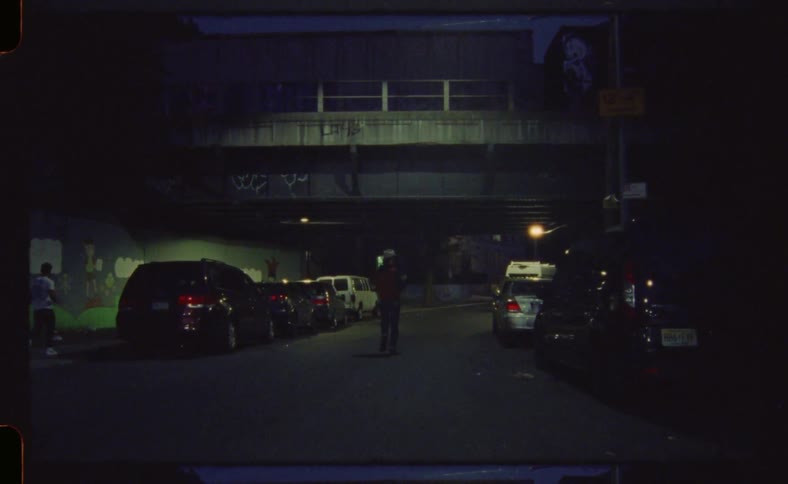 A man in a cowboy hat walks down the middle of the street at night with cars parked on the street