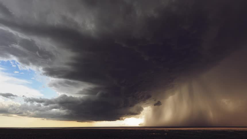 A large thunderstorm forming over a flat prairie with rain pouring down and the sun being hidden behind the strom
