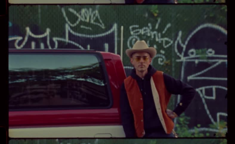 An old bronco truck parked in the street with a man wearing a hat and sunglasses leaning on the back of the truck during the day in the city