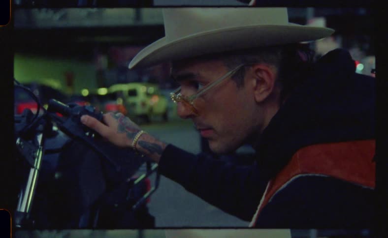 A man with tattooed hands and a cowboy hat is crouched down adjusting a part on a motorcycle parked on a city street at dusk