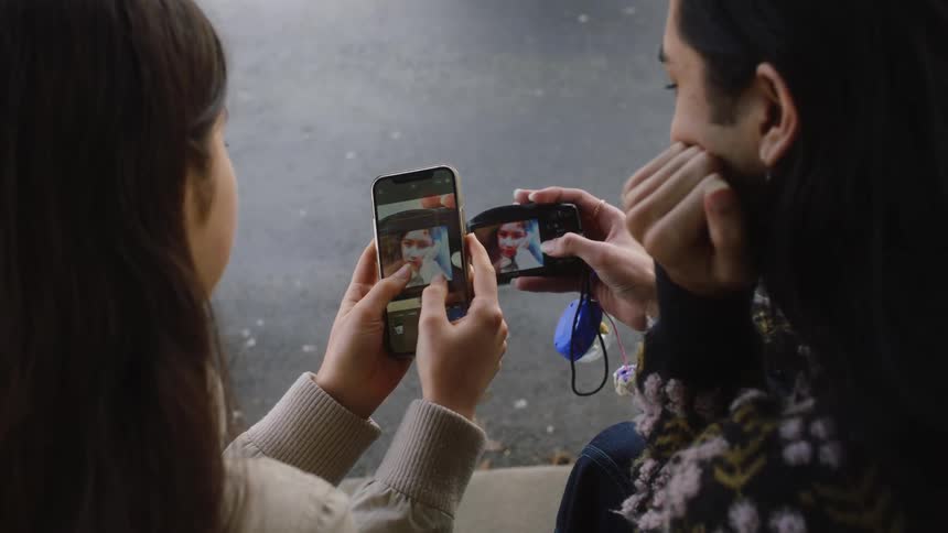 Two girls are taking pictures with a cell phone and a camera during the day