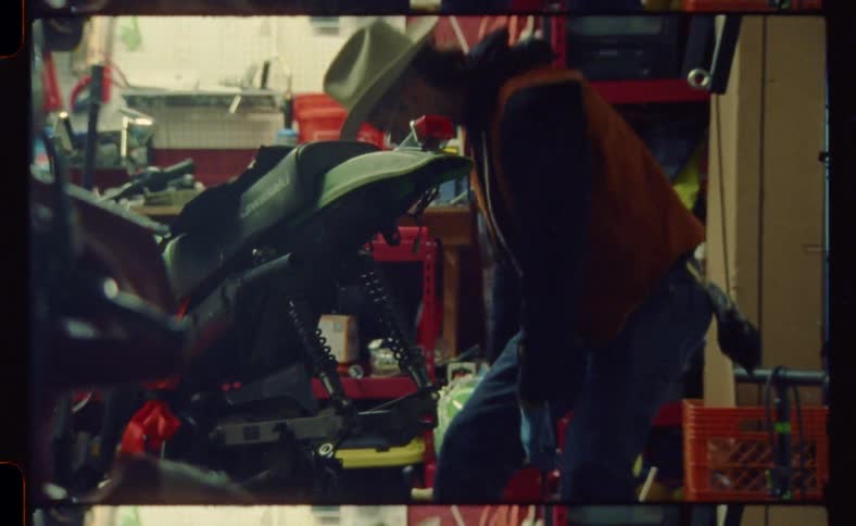 Man wearing a cowboy hat repairs a motorcycle in a small workshop