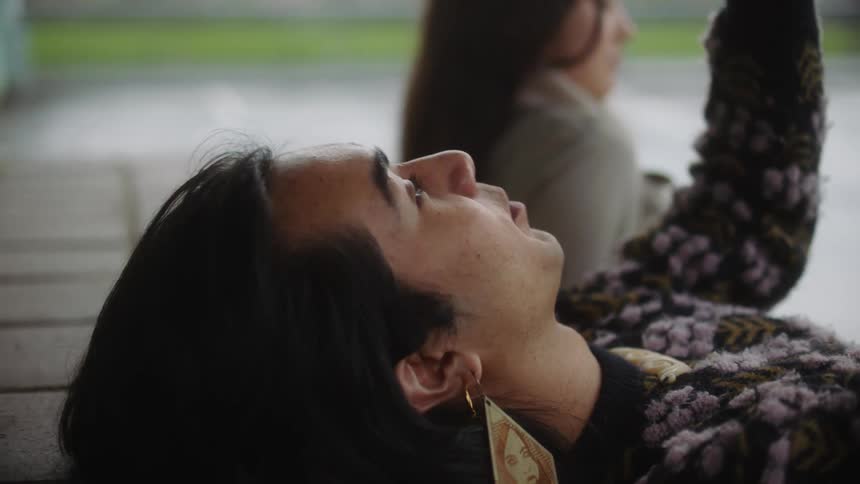 Man with long hair wearing a large earring talks to a friend while laying outdoors