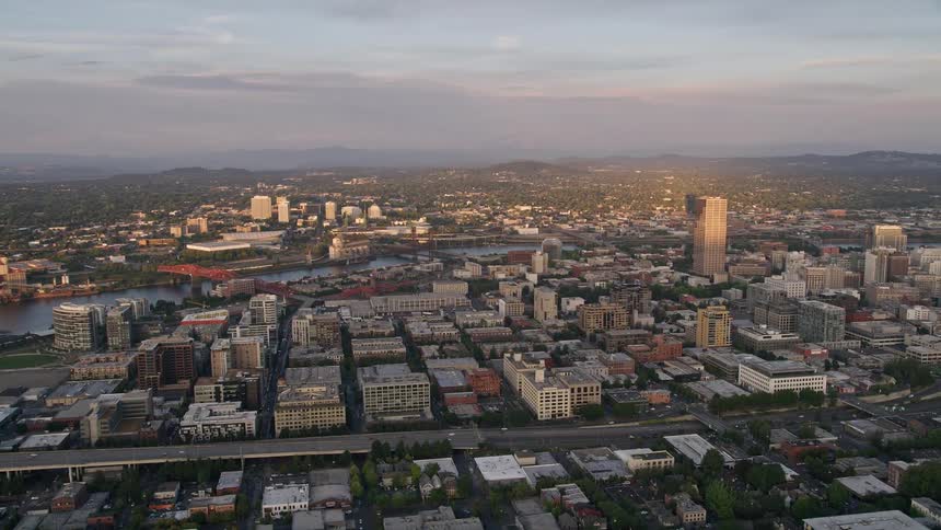 Aerial view showcasing downtown Portland's high-rise buildings the Lloyd District and the picturesque Willamette River at sunset with a partly cloudy sky adding to the charm - capturing the essence of this metropolis in the Pacific Northwest
