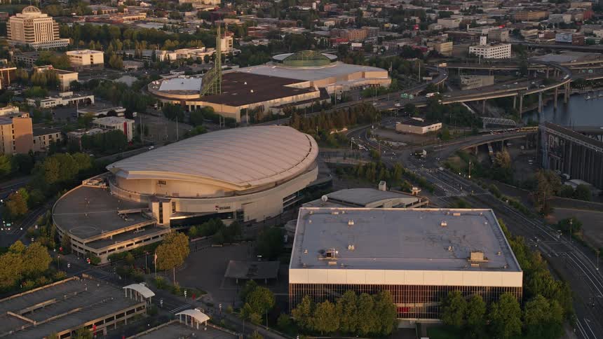 Drone footage captures the stunning sunset over the Rose Garden Arena and Memorial Coliseum in downtown Portland Oregon with the city's high-rise buildings and the river in the background