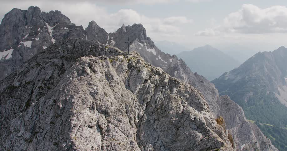 Aerial view of a mountain peak with a cross over it next to forest area and many other mountains during day time