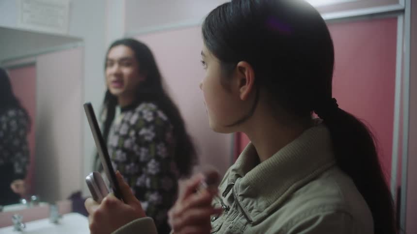 Two people doing their makeup one of them in front of the mirror and the other using a handheld accessory while talking at the same time