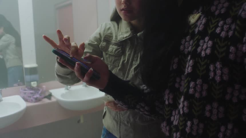 Two females use a cellphone standing in front of sinks and a mirror
