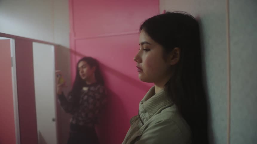 Two young women standing in a room leaning against the wall with the other one talking and one using her phone