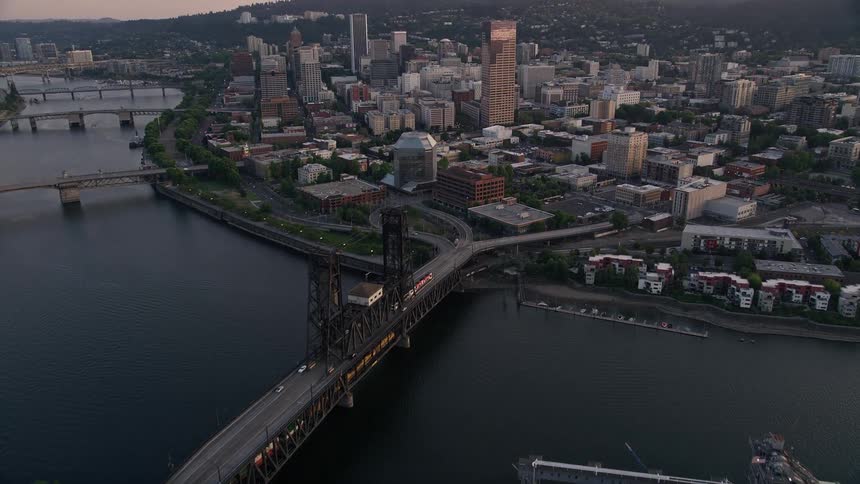 Aerial footage of downtown Portland Oregon at sunset capturing the Rose Garden Arena Steel Bridge and the city skyline with its high-rise buildings as well as the Willamette River and the lush nature surrounding it