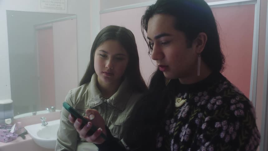 Two female friends in the bathroom talking to each other while standing and operating a mobile device