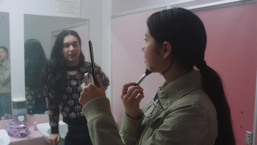 Two people in an indoor setting doing their makeup and talking to each other