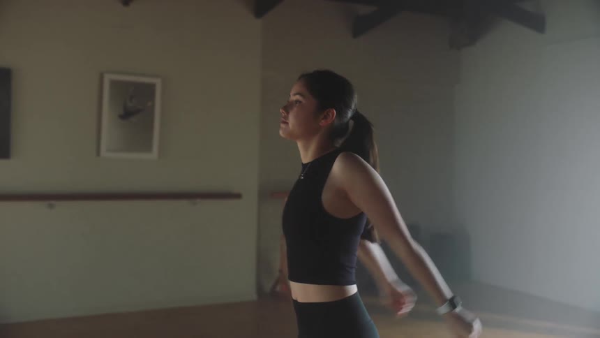 A young woman walking in a gymnasium before stopping and stretching while sun light comes in the windows
