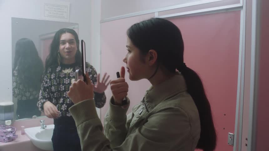 Two women in an indoor setting looking at themselves in the mirror and doing their makeup while having a conversation with each other