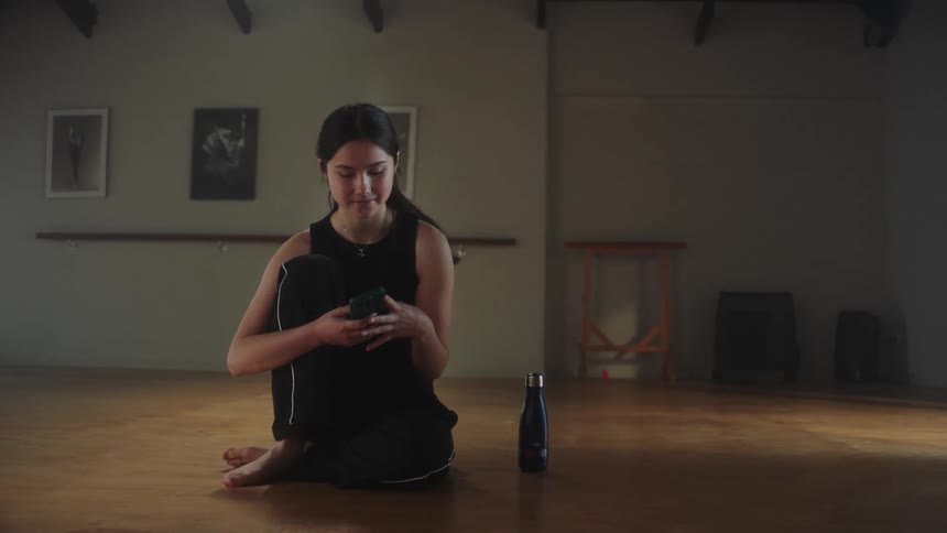 Female dancer sitting on dance studio floor checks smartphone and then leaves with her beverage