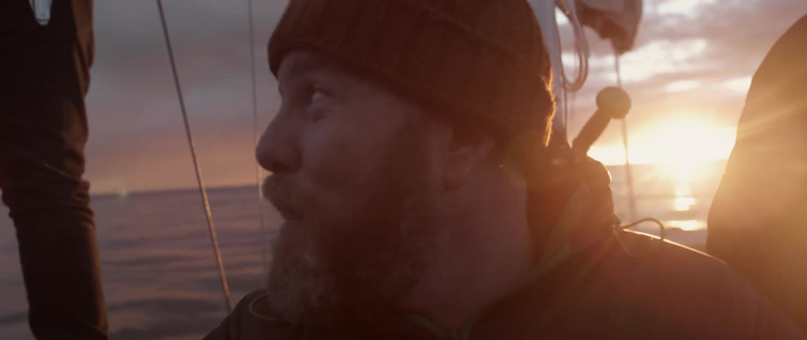 A man talks to someone next to him on a boat at dawn