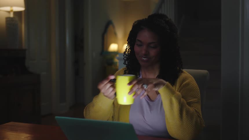A joyful black woman seated in the living room during the night happily checking something on her laptop she then picks up her yellow cup with a bright smile