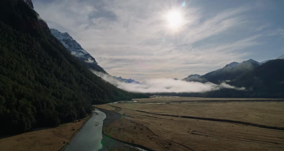 A landscape of a region where there is a river next to a forested hill and on the other side only undergrowth and in the sky the sun shining brightly