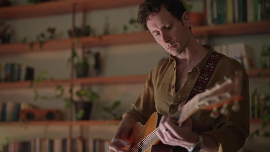 A man playing the guitar in a room filled with flower plant pots placed on shelves against the wall
