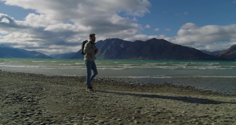 Man with a backpack walking alongside a beach surrounded by huge mountains and natural beauty