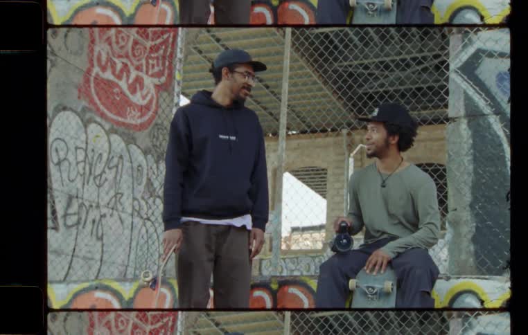 Two men with skateboards and camera in hand talking and laughing