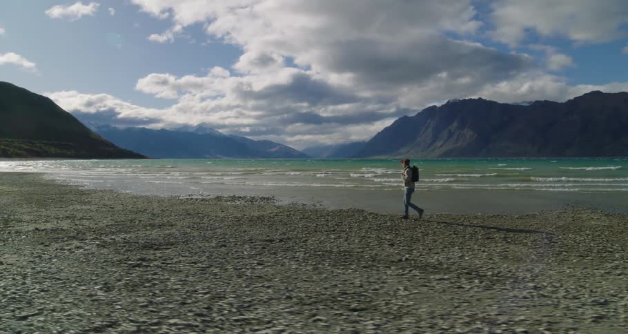 A man with a backpack is strolling along a rocky beach feeling the waves crash against the shore in the distance majestic mountains create a picturesque backdrop to the serene scene