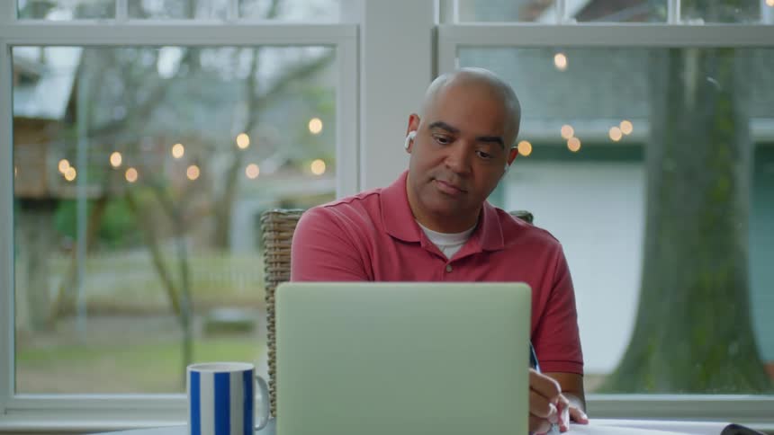 Man sitting looking at his laptop and jotting something down in a book with his pen