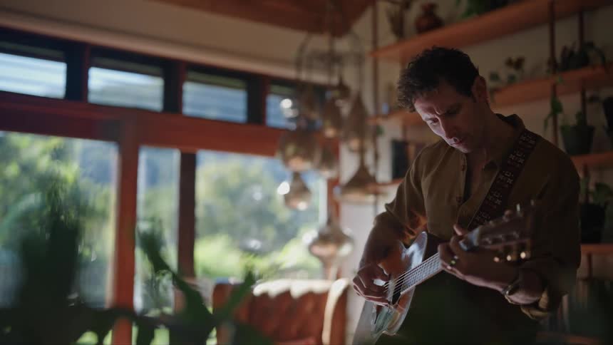 Man stands playing guitar in room with shelves full of plants