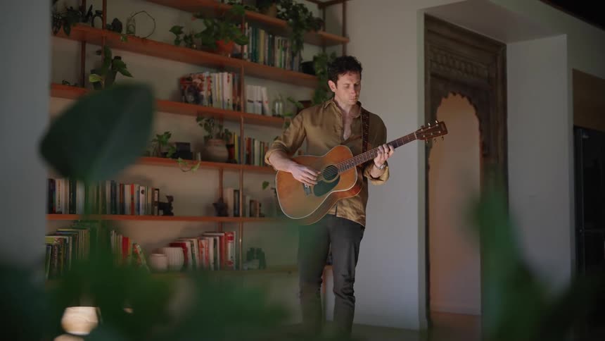 A man playing his guitar in his home in front of a book shelf during the day