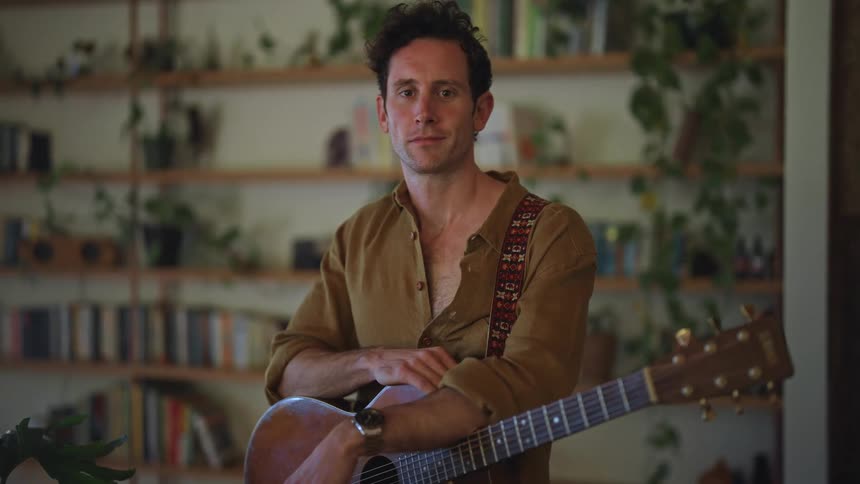 Man standing up in a room indoors holding a guitar and smiling during day time