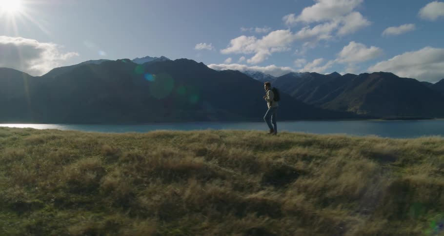 A man wearing a backpack is walking along the river bank with mountains in the distance
