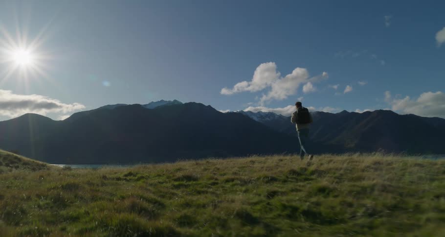 A man with a backpack walking on a lawn and behind him there is a huge river and mountains