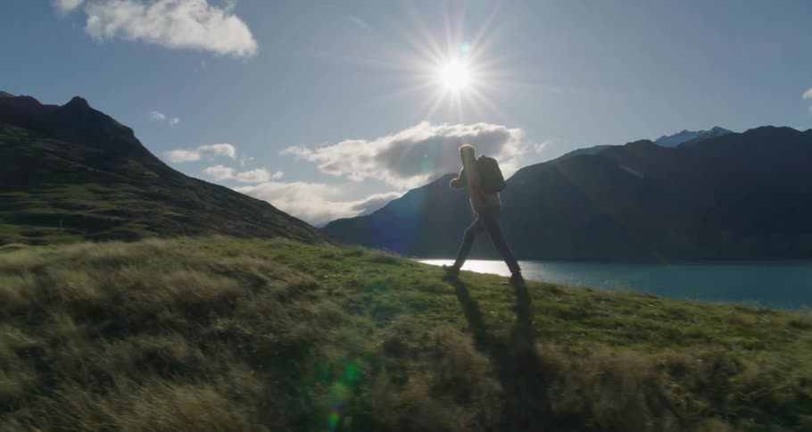 On a sunny day a man with a backpack hiking on a hill covered in grasses toward the mountain near a river