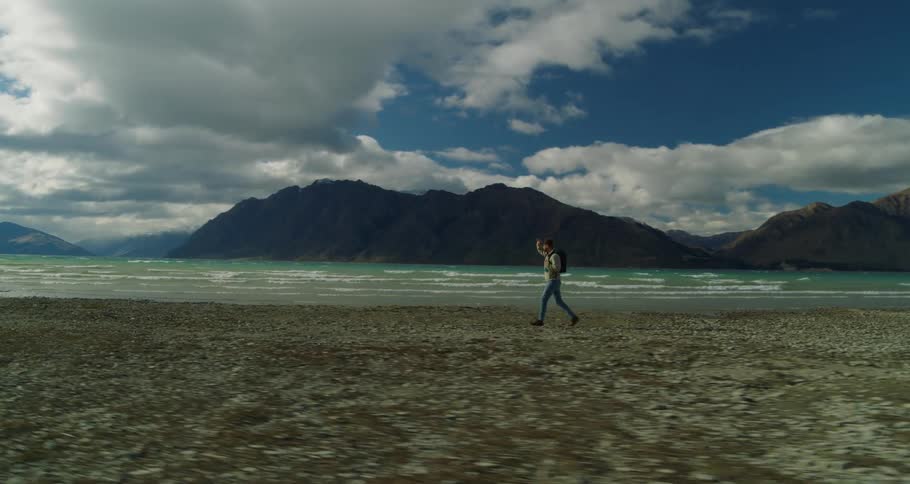 A hiker walks a line the shoreline next to a green watered lake opposite a low coastline mountain range under a blue sky with white clouds during the day
