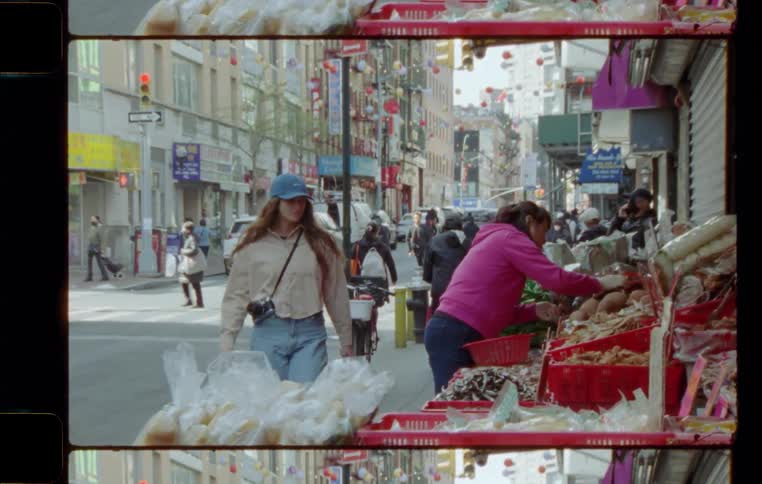 A woman tourist with a camera approaches food filled vender bins along a busy city sidewalk examining their contents