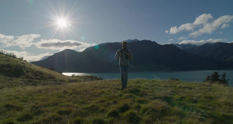 The sun shines upon a man wearing a backpack walking along the river bank with mountains in the distance