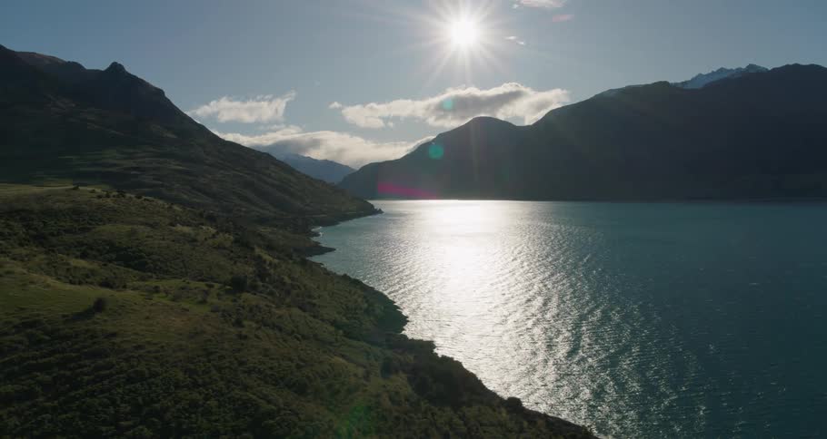 Aerial of the bright sun casting rays onto a body of water surrounded by grassy mountains