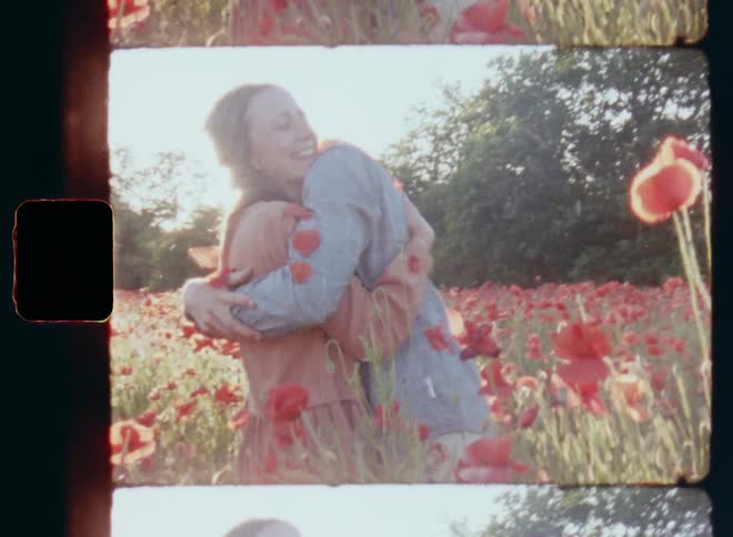 Young man of happy couple throws red petals of poppies in the air and hugs his girlfriend while both laugh smile and celebrate on a field with red flowers under intense sunlight on a warm summer evening on nostalgic 8mm film