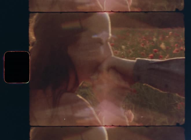 A young brown-haired woman smiles in a happy way and kisses the hand of her husband boyfriend or fiancé in a field of wildflowers on handheld dynamic 8mm film footage with bright sun light from the rear and retro lens flare