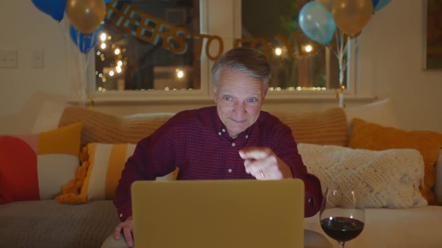 A man sitting on his couch with a glass of wine and a bowl of chips next to his laptop in front of him while he points to it