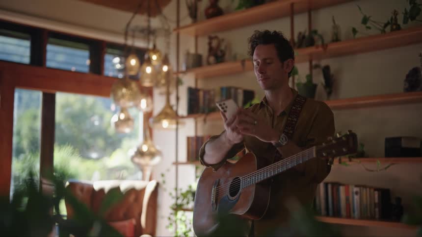 Young man playing acoustic guitar in the middle of the living room answers the phone