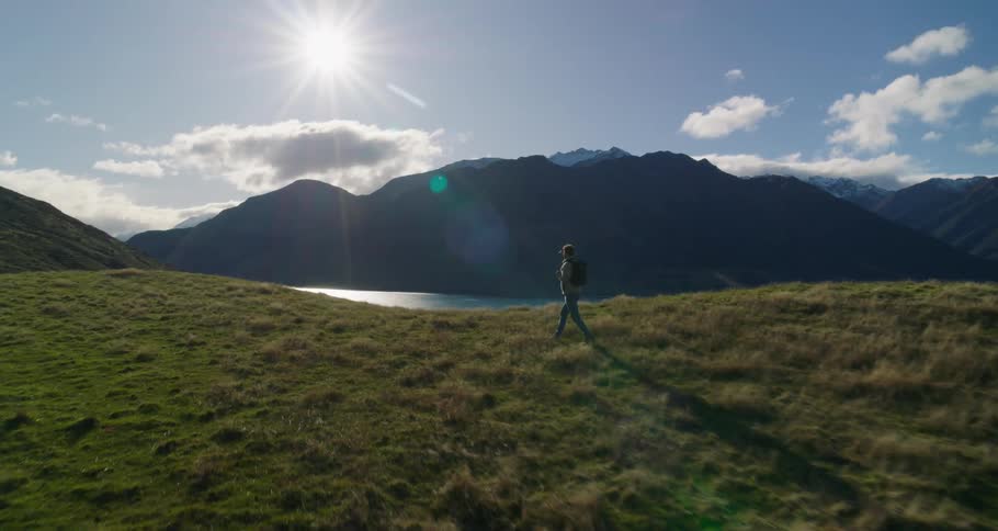 A man in a coat and backpack walking through a field of grass and right behind him is a huge river