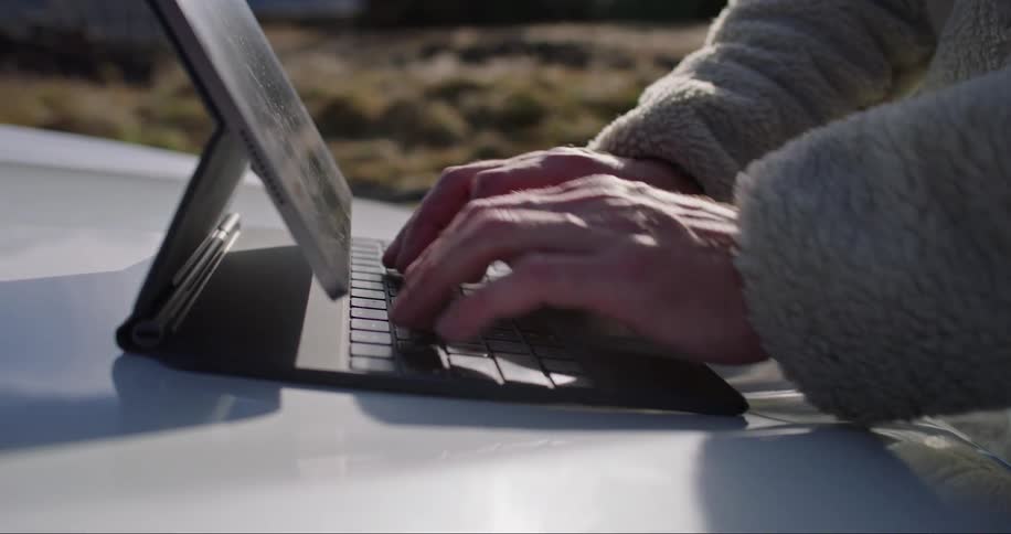 A person typing on a tablet pc while wearing a jacket with the tablet placed on top of a car trunk