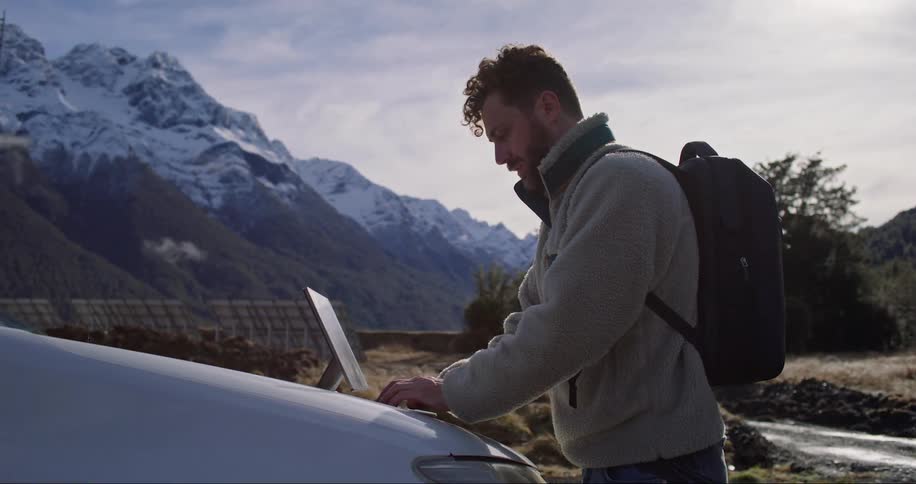 A man working on a notebook on top of the hood of a car and in the background there are huge mountains with ice on top