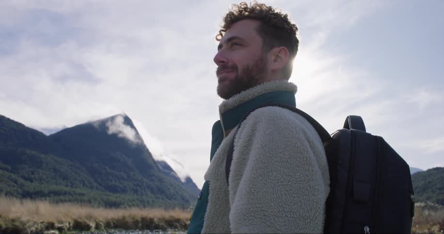 Young bearded man carrying a backpack stands on a valley near the mountains