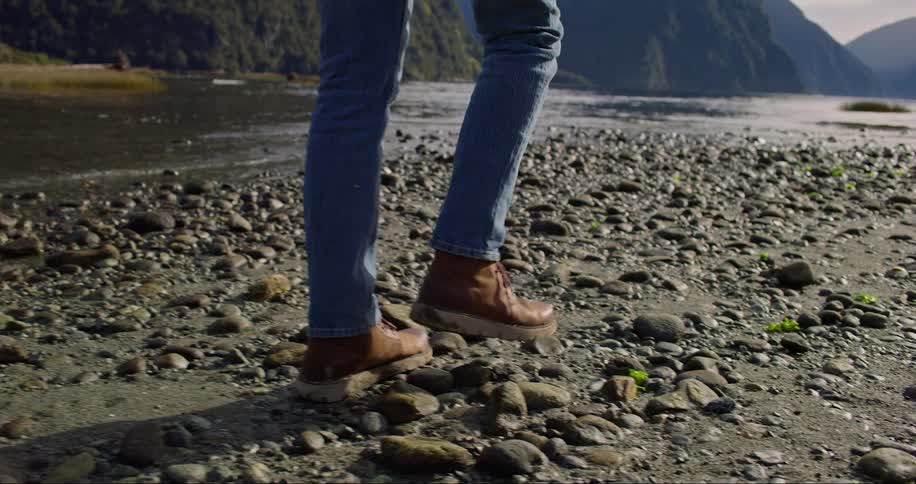 A man in blue jeans and brown work boots walking on a rocky lake shore next to mountains during the day
