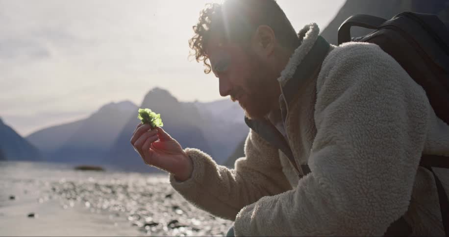 A man in a backpack and coat picks a leaf of moss out of the water and stares at it