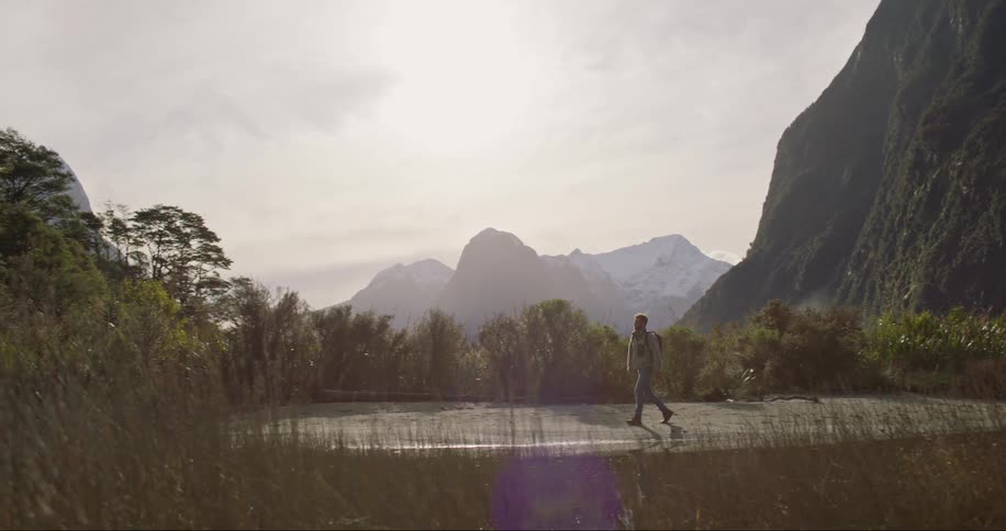 A man with a backpack is walking on a road in the middle of nature where in the background there are mountains and forest