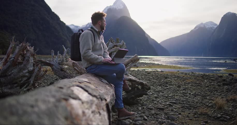 Man sitting using a laptop in a natural location with mountains and a lake